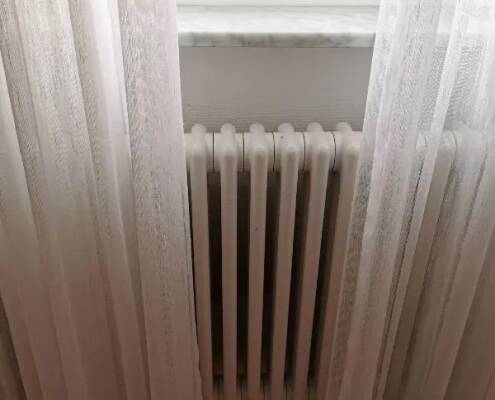 Cortinas largas blancas en velo sobre ventana posicionadas sobre un radiador en hierro fundido blanco