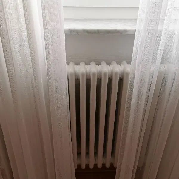 Cortinas largas blancas en velo sobre ventana posicionadas sobre un radiador en hierro fundido blanco